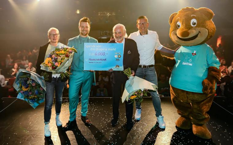 4 Personen und ein übergrosses Maskottchen stehen auf einer Bühne mit einem großen Scheck. Im Hintergrund ein voller Saal.
